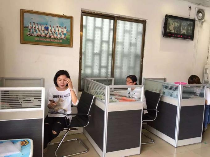 Office space with employees working at cubicles, framed team photo on wall, digital clock, and window with frosted glass panels — reflecting daily administrative operations.